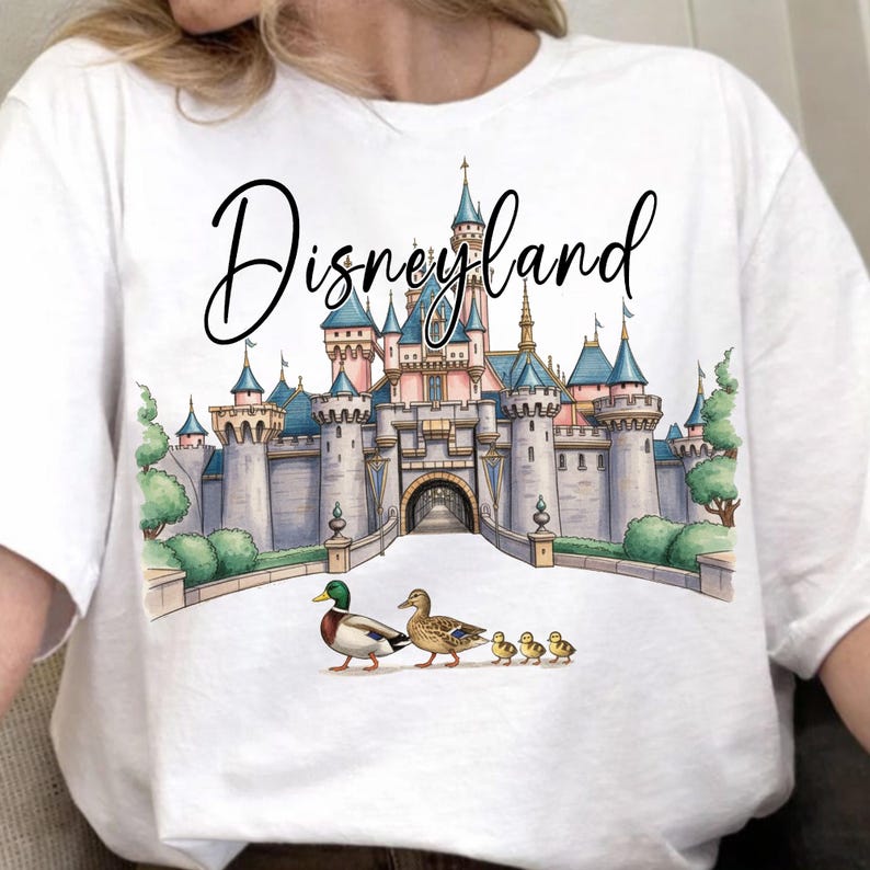 May include: White t-shirt featuring a watercolor illustration of Disneyland's castle with ducks. The word "Disneyland" is written in a cursive font above the castle. The ducks are walking in a line.