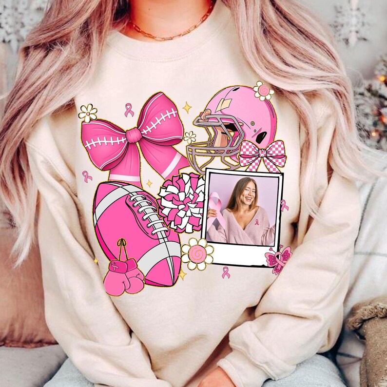 May include: A beige sweatshirt featuring pink football-themed graphics, including a football, helmet, bow, and boxing gloves. A photo of a woman holding a pink ribbon is also included. Pink ribbons and flowers are scattered around the design.