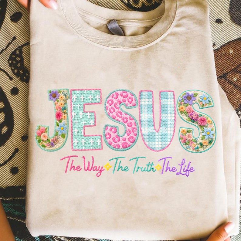 Puede incluir: Camiseta beige con la palabra "JESUS" en un dise&ntilde;o bordado colorido. Las letras presentan patrones florales, de leopardo y a cuadros. Debajo, el texto dice "The Way The Truth The Life" en una fuente de escritura.