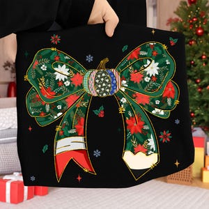 May include: Black decorative bow with a festive design. The bow features a pumpkin-shaped center with various patterns and a green ribbon adorned with red poinsettias, white flowers, and gold accents. The bow is held up by a person.