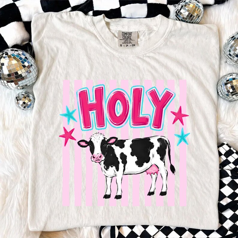Puede incluir: Camiseta color crema con un dise&ntilde;o de una vaca en blanco y negro con ubres rosas. La palabra "HOLY" en rosa con contorno azul, estrellas rosas y azules, sobre un fondo de rayas rosas.
