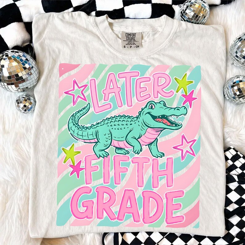 May include: Cream-colored t-shirt featuring a graphic of a teal alligator with the words "LATER FIFTH GRADE" in pink. The design includes pink and green stars and stripes. The shirt is a Comfort Colors brand.