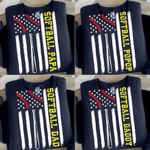 Op de afbeelding: Marineblauwe sweatshirts met een ontwerp dat lijkt op de Amerikaanse vlag, met een softbal en een knuppel. De tekst op de sweatshirts luidt "SOFTBALL PAPA", "SOFTBALL POP", "SOFTBALL DAD" en "SOFTBALL DADDY" in het geel.