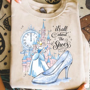 May include: Beige t-shirt featuring a Cinderella-themed design. The graphic includes a clock, a castle, Cinderella in a blue gown, and a glass slipper. Text reads "it's all about the Shoes."