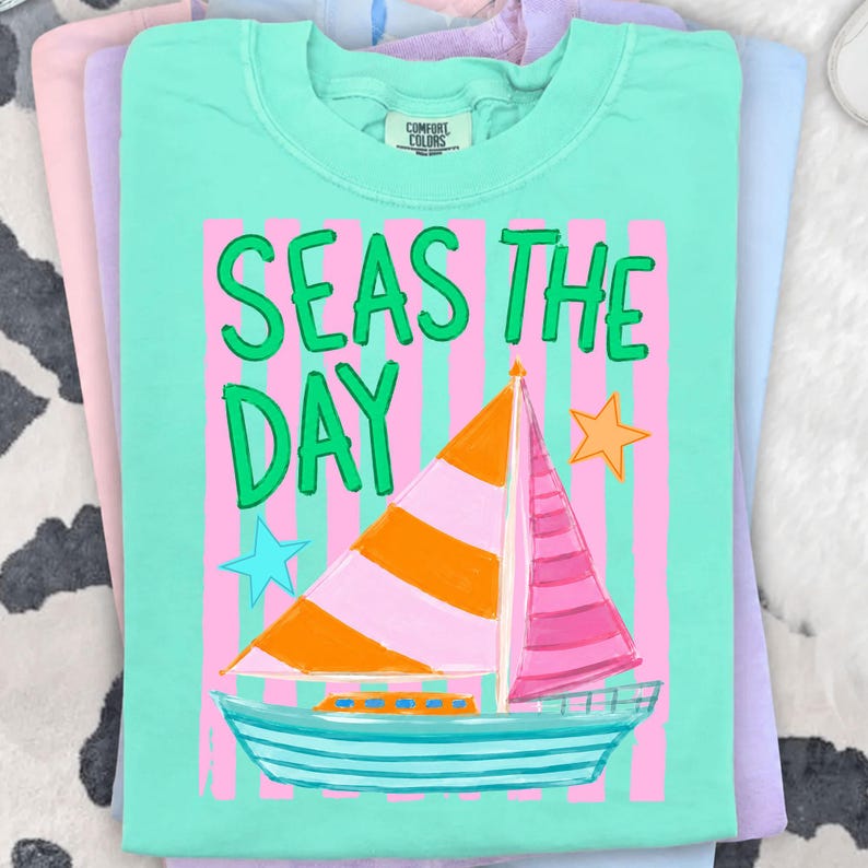 May include: A turquoise Comfort Colors t-shirt with the phrase "SEAS THE DAY" in green lettering. The shirt features a sailboat graphic with orange and pink sails, blue and yellow stars, and pink vertical stripes.
