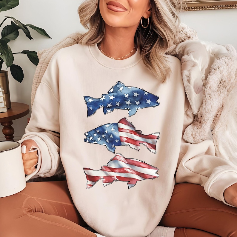 May include: A cream-coloured sweatshirt with three fish designs. The top fish is blue with white stars, the middle fish has a US flag design, and the bottom fish is red and white striped. The sweatshirt is a casual style.