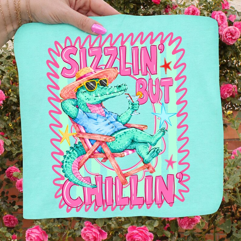 May include: A turquoise tote bag featuring a cartoon alligator in a beach chair, wearing sunglasses and a hat, holding a cocktail. The text "Sizzlin' But Chillin'" is in pink, with a pink wavy border.