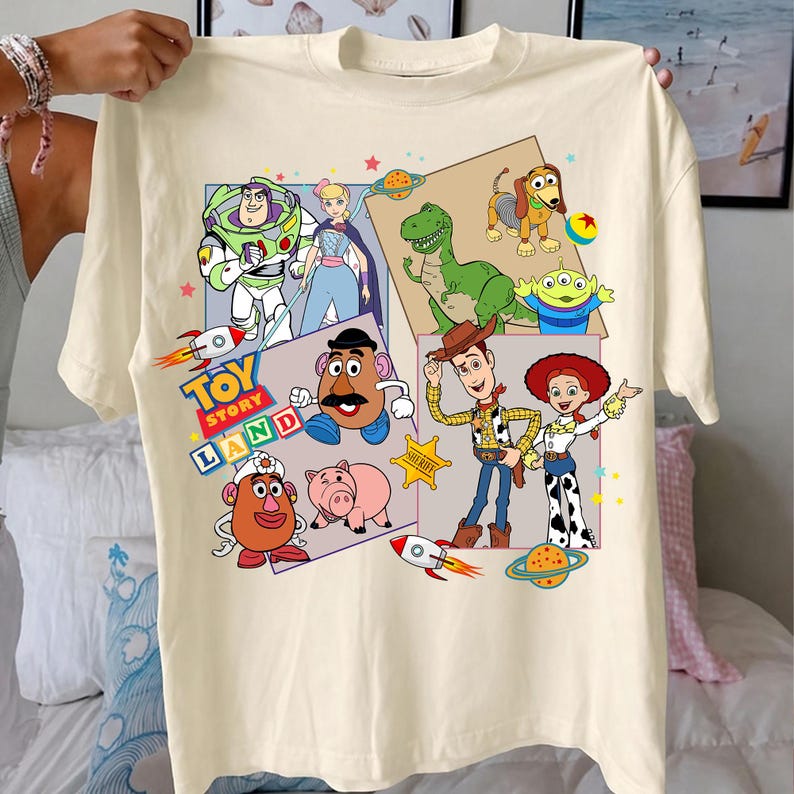 Puede incluir: Camiseta de color crema con un gr&aacute;fico colorido de los personajes de Toy Story, incluyendo a Buzz Lightyear, Woody y Jessie. El dise&ntilde;o incluye el texto "Toy Story Land" con cohetes y planetas.