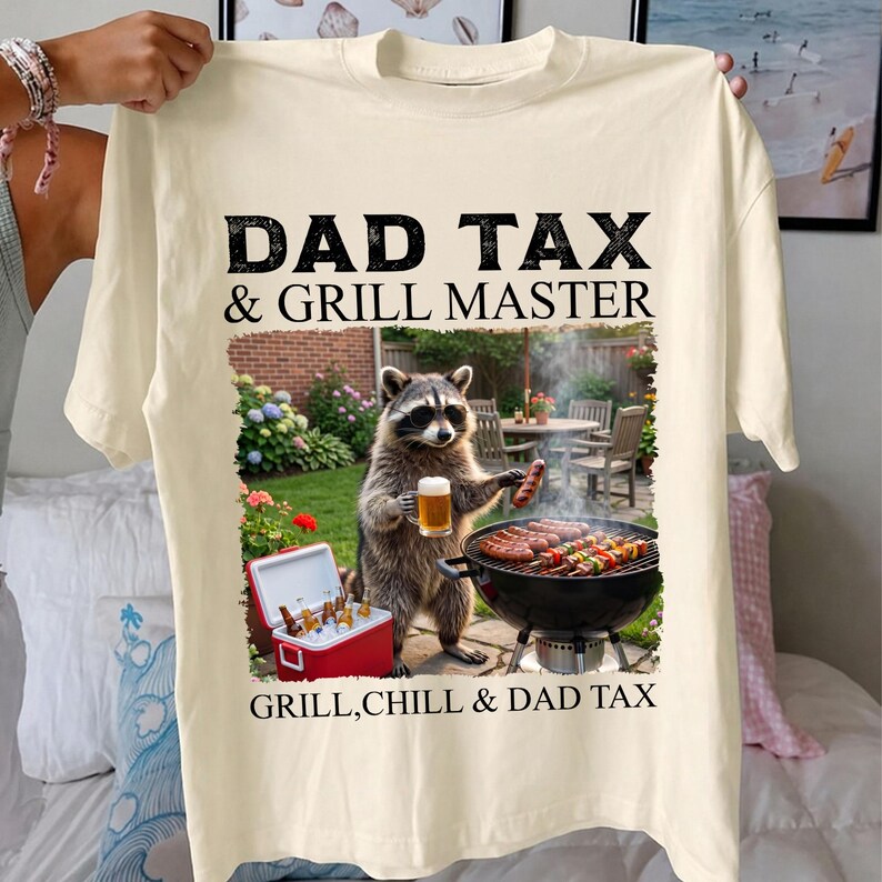 May include: Cream-colored t-shirt with the text "DAD TAX & GRILL MASTER" in black. A raccoon graphic holds a beer and sausage near a grill. The text "GRILL, CHILL & DAD TAX" is below the graphic.