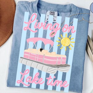 May include: Light blue t-shirt with a graphic of a pink and white pontoon boat, a yellow sun, and the words "Living on Lake time" in pink script. The shirt has a Comfort Colors tag.