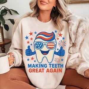 May include: Cream-colored sweatshirt featuring a cartoon tooth with sunglasses, thumbs up, and a patriotic design. The text "MAKING TEETH GREAT AGAIN" is printed below the tooth in red and blue. Stars and fireworks surround the tooth.