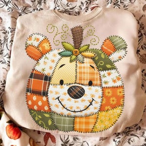 May include: A light pink sweatshirt featuring a patchwork pumpkin design. The pumpkin is made of various fabric patterns in orange, yellow, and green, with a smiling face and a stem. The design is stitched onto the sweatshirt.