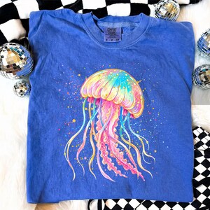 May include: A blue Comfort Colors sweatshirt featuring a colorful jellyfish design. The jellyfish is depicted with a rainbow of colors, including pink, yellow, blue, and green. The sweatshirt has a crew neck and the Comfort Colors logo is visible.