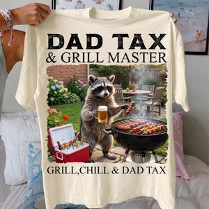 May include: Cream-colored t-shirt with the text "DAD TAX & GRILL MASTER" in black. The graphic features a raccoon wearing sunglasses, holding a beer and a sausage, grilling. A cooler with bottles is also visible.