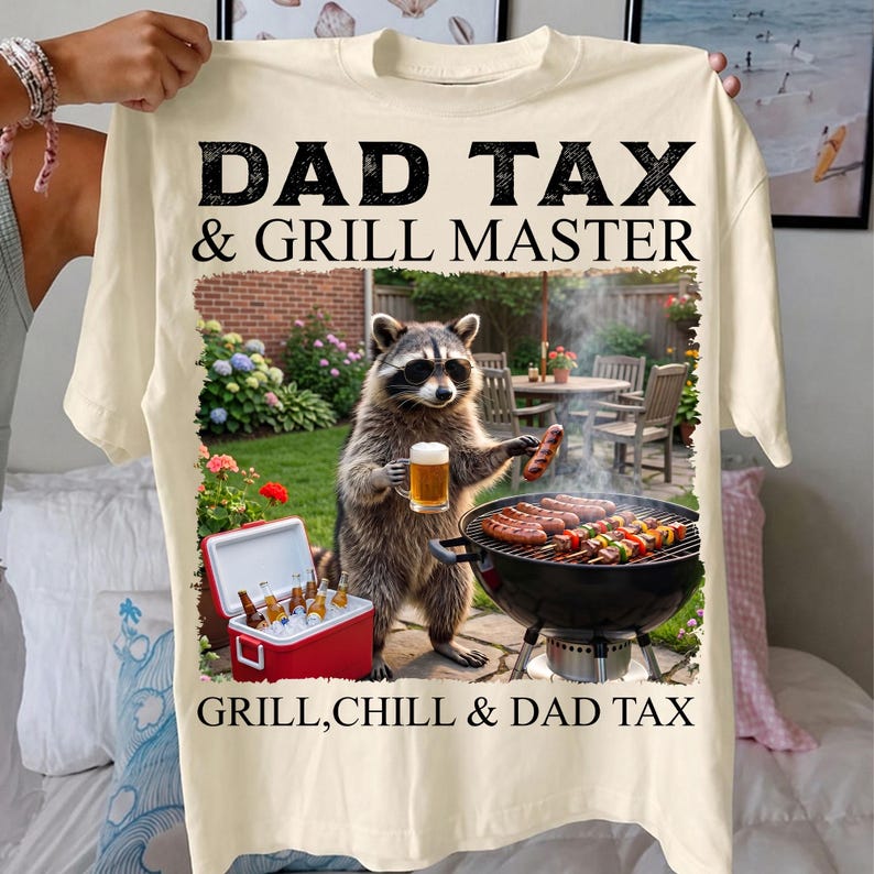 May include: Cream-colored t-shirt with the text "DAD TAX & GRILL MASTER" in black. The graphic features a raccoon wearing sunglasses, holding a beer and a sausage, grilling. A cooler with bottles is also visible.