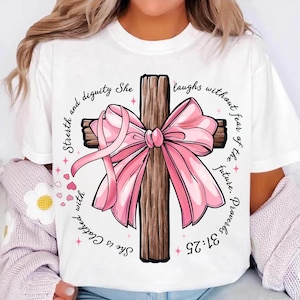 May include: White t-shirt with a wooden cross design, embellished with a pink ribbon and a pink ribbon-shaped breast cancer awareness symbol. The design includes the text "She is Clothed with Strength and Dignity" and "Proverbs 31:25."