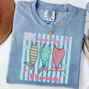 May include: A light blue Comfort Colors t-shirt with a graphic that says "Wanna Play Mermaids!" The graphic features three mermaid tails in red, green, and pink, hanging on a clothesline with starfish and bows.