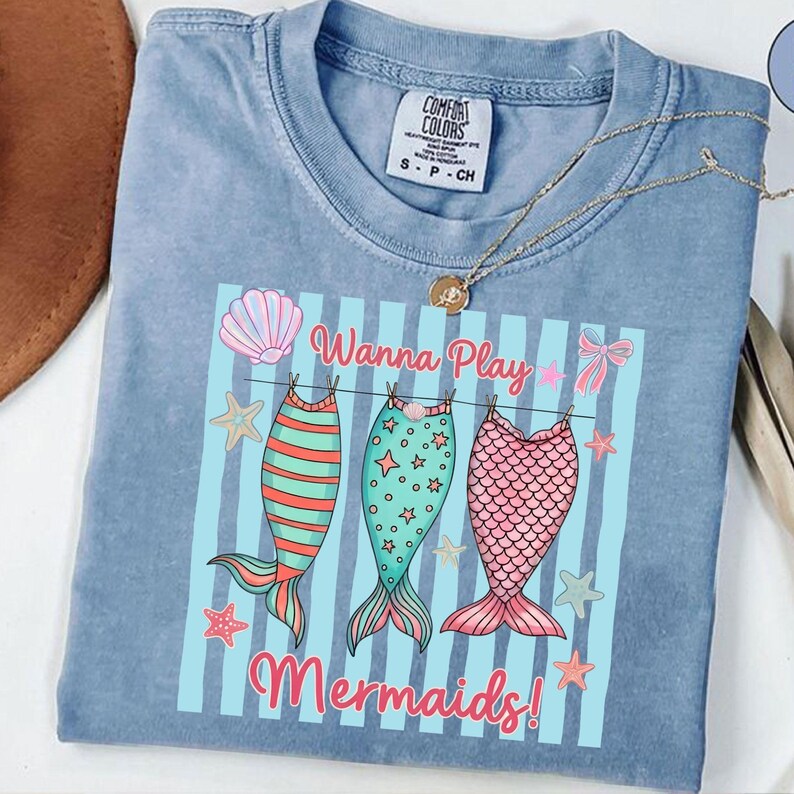May include: A light blue Comfort Colors t-shirt with a graphic that says "Wanna Play Mermaids!" The graphic features three mermaid tails in red, green, and pink, hanging on a clothesline with starfish and bows.