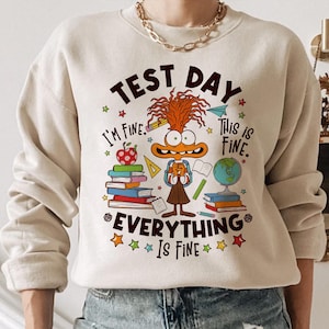 May include: A beige sweatshirt with a cartoon character wearing a backpack and holding books. The text on the sweatshirt reads "Test Day I'm Fine. This is Fine. Everything is Fine."