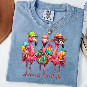 May include: Light blue Comfort Colors t-shirt featuring a graphic of three pink flamingos. One flamingo wears a green cap and holds an ice cream cone. Another wears a Hawaiian shirt and holds a popsicle. The third wears sunglasses and a straw hat, holding a drink.