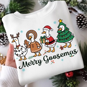 Peut inclure: Sweat-shirt blanc avec un motif festif repr&eacute;sentant des oies de dessin anim&eacute; v&ecirc;tues de tenues de f&ecirc;te. Une oie porte des bois de renne, une autre tient un bonhomme en pain d'&eacute;pices, et d'autres sont en costumes de P&egrave;re No&euml;l et de sapin de No&euml;l. Le texte "Merry Goosmas" est imprim&eacute; en dessous.