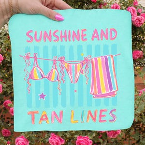 Peut inclure: Une serviette de plage turquoise avec les mots "SUNSHINE AND TAN LINES" en rose et jaune. La serviette repr&eacute;sente un bikini et une serviette ray&eacute;e suspendus &agrave; une corde &agrave; linge. Une &eacute;toile est en dessous.
