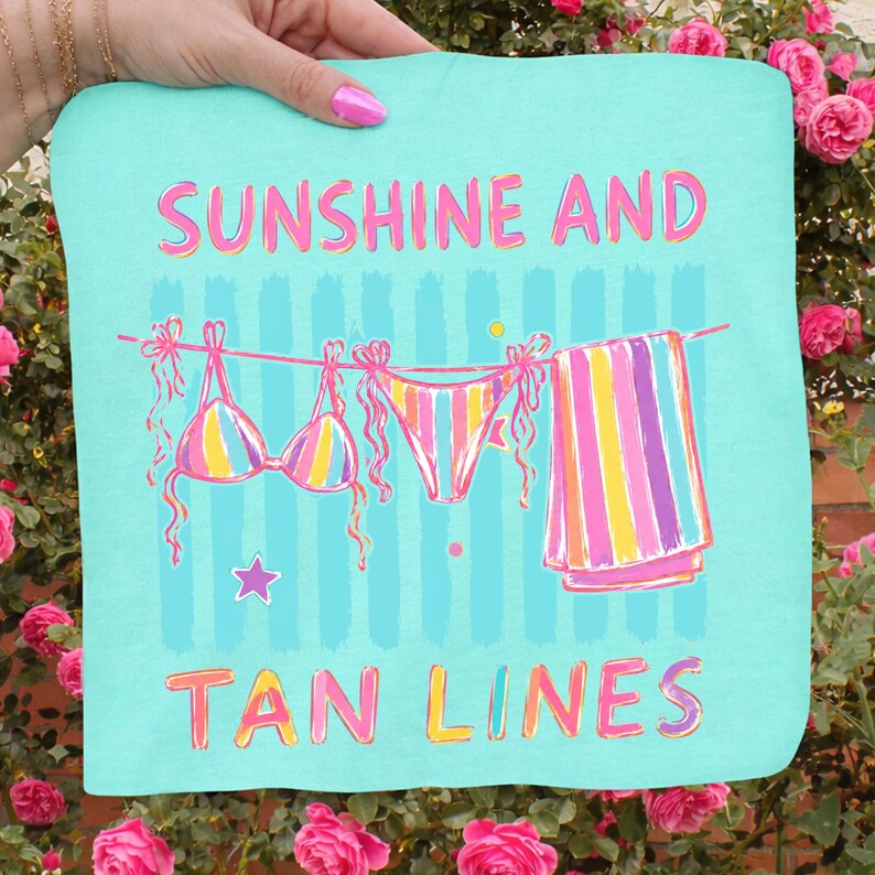 Peut inclure: Une serviette de plage turquoise avec les mots "SUNSHINE AND TAN LINES" en rose et jaune. La serviette repr&eacute;sente un bikini et une serviette ray&eacute;e suspendus &agrave; une corde &agrave; linge. Une &eacute;toile est en dessous.