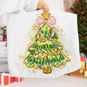 May include: A white Christmas-themed pillow featuring a colorful, hand-drawn Christmas tree design. The tree is decorated with a pink bow, gold star, and candy canes. The background has a splatter effect with gold and green accents.