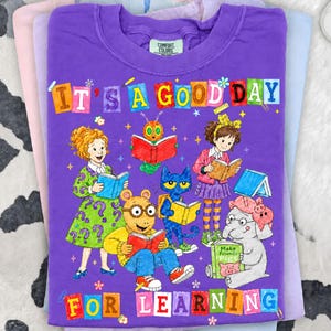 Puede incluir: Una camiseta morada de manga corta con un gr&aacute;fico colorido de personajes de dibujos animados leyendo libros. El texto dice IT'S A GOOD DAY FOR LEARNING. El dise&ntilde;o incluye una ni&ntilde;a, una oruga, un gato, un cerdo hormiguero y un elefante.