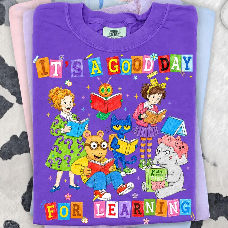 Puede incluir: Una camiseta morada de manga corta con un gr&aacute;fico colorido de personajes de dibujos animados leyendo libros. El texto dice IT'S A GOOD DAY FOR LEARNING. El dise&ntilde;o incluye una ni&ntilde;a, una oruga, un gato, un cerdo hormiguero y un elefante.