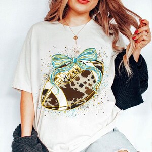 May include: Cream-colored t-shirt featuring a football design with a cowhide pattern and a blue bow. The football has gold accents and white stripes. The shirt is a casual style.