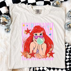 May include: White t-shirt with a cartoon mermaid graphic. The mermaid has red hair, heart-shaped sunglasses, and a bubble. The design is set against a pink and white striped background with starfish and seashells. The shirt has a Comfort Colors tag.