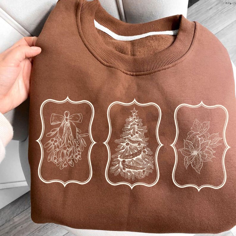 May include: A brown sweatshirt featuring three white framed holiday designs: mistletoe, a Christmas tree, and poinsettias. The sweatshirt has a crew neck and a white stripe on the inside of the collar.