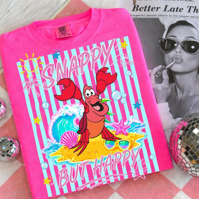 May include: Hot pink t-shirt with a graphic of a cartoon crab, sunglasses, and the words "Snappy But Happy." The design includes a wave, palm trees, and a starfish against a striped background.