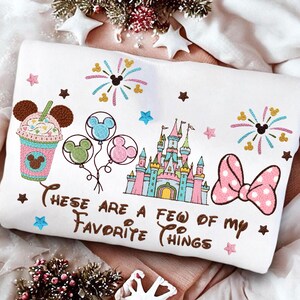 May include: White sweatshirt with colorful embroidered Disney-themed graphics. Features a castle, balloons, a drink, fireworks, and a bow. Text reads "These are a few of my favorite things."