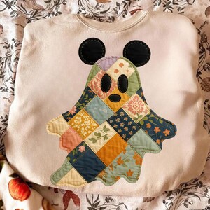 May include: Cream-colored sweatshirt featuring a patchwork ghost design with Mickey Mouse ears. The ghost is made of various floral and patterned fabrics in shades of blue, orange, pink, and green. Black circular ears are stitched on top.