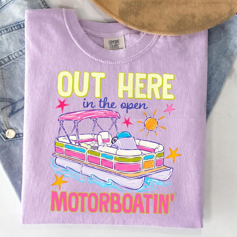 May include: A lilac Comfort Colors t-shirt with a colorful graphic of a pontoon boat. The text "OUT HERE in the open MOTORBOATIN'" is printed in yellow, pink, and blue. The design includes a sun and stars.