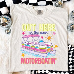May include: Cream-colored t-shirt with a colorful graphic of a pontoon boat and the text "OUT HERE in the open MOTORBOATIN'". The boat has a pink canopy and is surrounded by stars and a sun.