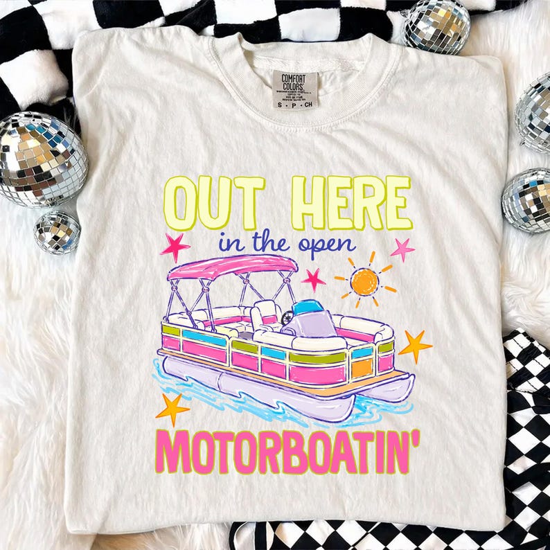 May include: Cream-colored t-shirt with a colorful graphic of a pontoon boat and the text "OUT HERE in the open MOTORBOATIN'". The boat has a pink canopy and is surrounded by stars and a sun.