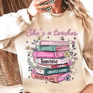 Op de afbeelding: Beige sweatshirt met de roze tekst "She's a teacher". Het ontwerp toont een stapel boeken met bloemversieringen en de woorden "Strong", "Victorious", "Beautiful", "Capable", "Greatly Loved" en "Never Alone".
