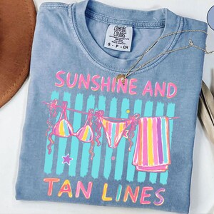 Peut inclure: T-shirt bleu clair Comfort Colors avec les mots "SUNSHINE AND TAN LINES" en rose et jaune. Le t-shirt pr&eacute;sente un motif de bikini, de sous-v&ecirc;tements et d'une serviette suspendus &agrave; une corde &agrave; linge. Un collier dor&eacute; est drap&eacute; sur le t-shirt.