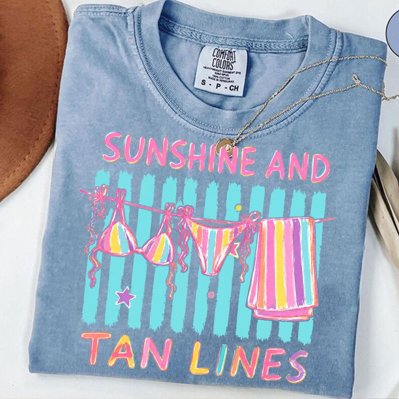 Peut inclure: T-shirt bleu clair Comfort Colors avec les mots "SUNSHINE AND TAN LINES" en rose et jaune. Le t-shirt pr&eacute;sente un motif de bikini, de sous-v&ecirc;tements et d'une serviette suspendus &agrave; une corde &agrave; linge. Un collier dor&eacute; est drap&eacute; sur le t-shirt.