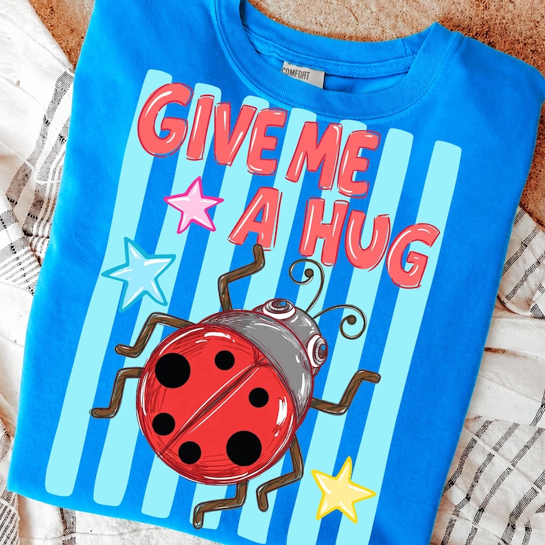 Pu&ograve; includere: Una maglietta blu con la scritta "GIVE ME A HUG" in lettere rosse. La maglietta presenta una coccinella dei cartoni animati con un corpo rosso e macchie nere, insieme a grafiche a stella. La maglietta ha strisce verticali blu e bianche.