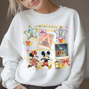 May include: White sweatshirt featuring a colorful graphic of Disney characters, including Mickey and Minnie Mouse, Donald and Daisy Duck, and Goofy with Pluto. The word "Magical" is written above the images.