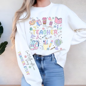 May include: White sweatshirt with colorful "TEACHER" graphic and school-themed illustrations. Features a coffee cup, apple, globe, and backpack. The left sleeve has text that reads "Can't talk right now Doing TEACHER STUFF".