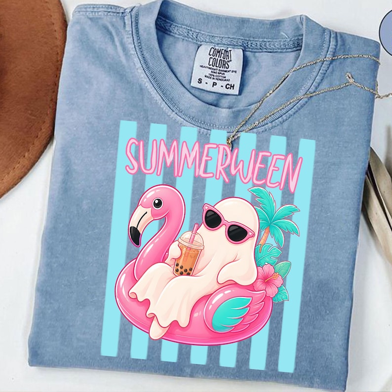 K&ouml;nnte beinhalten: Hellblaues T-Shirt mit einer Grafik eines Geistes mit Sonnenbrille, der auf einem rosa Flamingo-Schwimmring schwimmt und ein Getr&auml;nk trinkt. Das Wort "SUMMERWEEN" ist in rosa Buchstaben &uuml;ber der Grafik aufgedruckt.