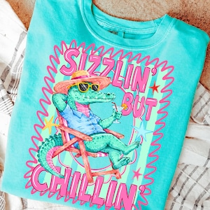 May include: Turquoise crew neck sweatshirt with a graphic of an alligator wearing sunglasses and a straw hat, relaxing in a beach chair and holding a cocktail. The text reads "Sizzlin' But Chillin'" in pink lettering.