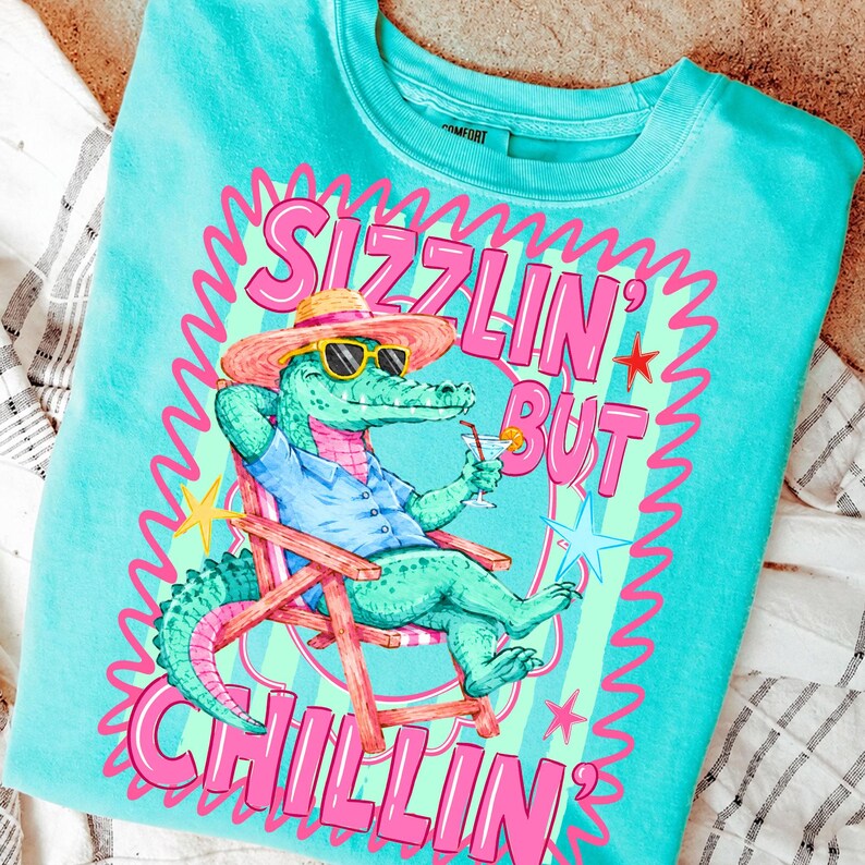 May include: Turquoise crew neck sweatshirt with a graphic of an alligator wearing sunglasses and a straw hat, relaxing in a beach chair and holding a cocktail. The text reads "Sizzlin' But Chillin'" in pink lettering.