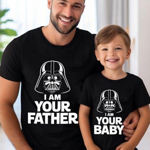 May include: Black t-shirts with white graphic of Darth Vader's helmet. One shirt reads "I AM YOUR FATHER", the other reads "I AM YOUR BABY". The shirts are worn by a father and child.