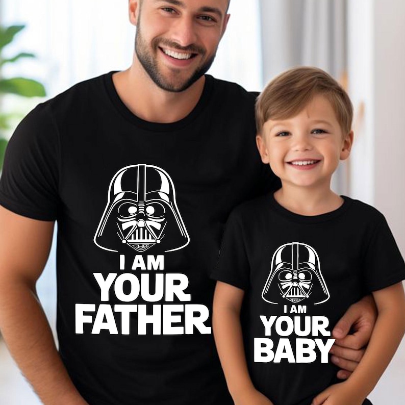 May include: Black t-shirts with white graphic of Darth Vader's helmet. One shirt reads "I AM YOUR FATHER", the other reads "I AM YOUR BABY". The shirts are worn by a father and child.